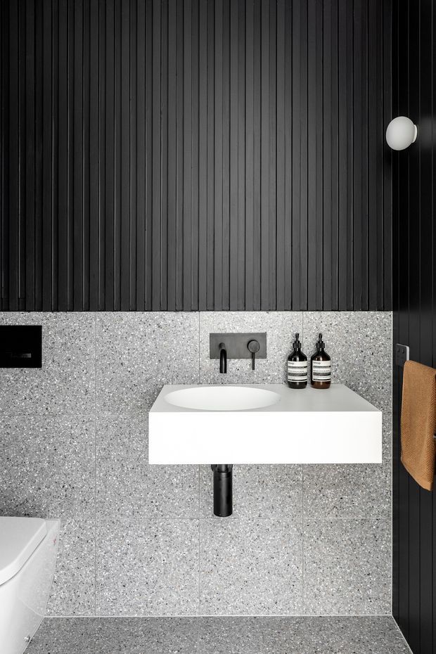 A compact contemporary bathroom featuring a wall-mounted white vanity with an integrated round basin, black wall-mounted tapware and exposed drain, grey terrazzo wall and floor tiles, and black vertically ribbed wall panelling. A partial toilet, black flush plate, round wall light, and tan hand towel are visible.