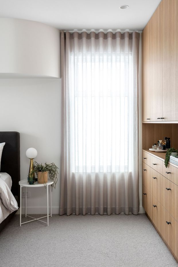 Bright, compact bedroom corner with a tall sheer-curtained window, taupe drapes, light grey carpet, pale walls, and a full-height built-in timber wardrobe and dressing cabinet. A dark upholstered bed edge, small round bedside table, globe lamp, and potted plant are visible. The room has a clean, calm, contemporary feel with warm wood joinery and abundant natural light.