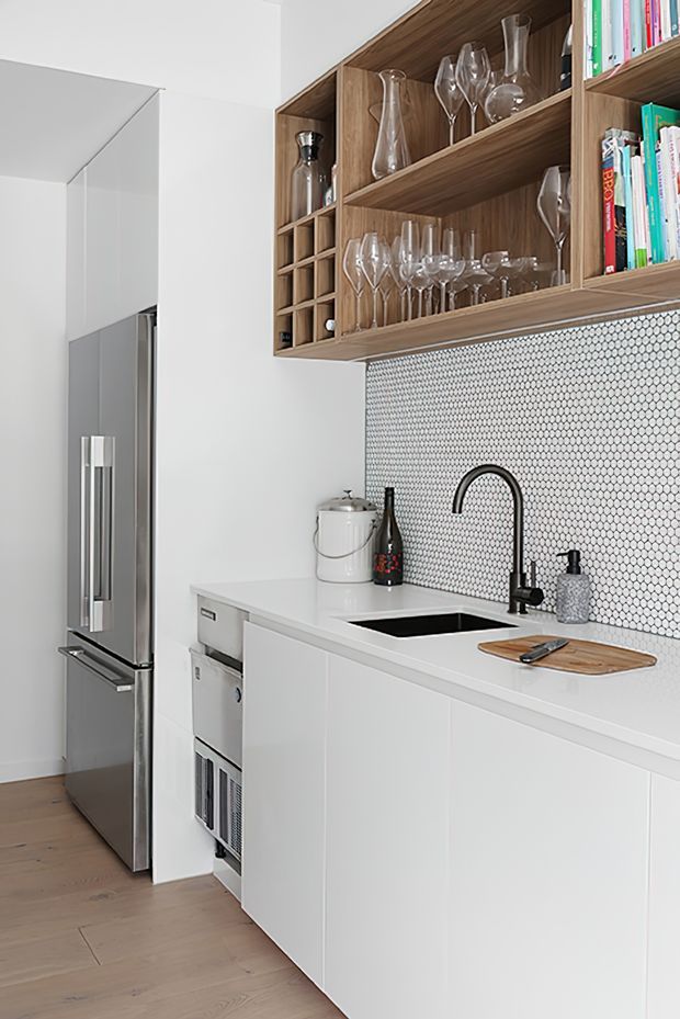 Bright, compact contemporary kitchen with white handleless base cabinetry, a white benchtop, black undermount sink and curved black tap. A small-scale white and grey hexagonal mosaic splashback sits beneath open walnut-toned shelving displaying wine glasses, bottles and books. Stainless-steel refrigeration and integrated appliances are visible at left, with pale timber flooring throughout.