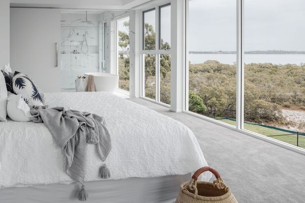 Bright contemporary bedroom with a white upholstered bed, textured white bedding, grey throw, and patterned accent cushions. Floor-to-ceiling corner glazing provides expansive views over treetops and a waterway, while a freestanding white bathtub and glass shower are visible behind the bed, creating a bedroom-bathroom suite. The space has pale carpet, white walls, light-toned finishes, and abundant natural light.