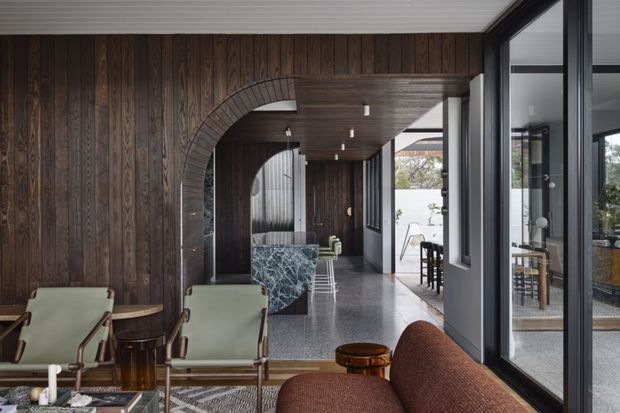 A sophisticated open-plan interior with a dark timber-lined feature wall and ceiling, large arched opening, polished terrazzo-style flooring, and expansive black-framed glazing. The foreground contains mid-century-inspired lounge seating and small tables, while a marble-patterned island or bar and dining area extend through the connected space. The palette is dark, warm, and refined, with strong indoor-outdoor visual connection.
