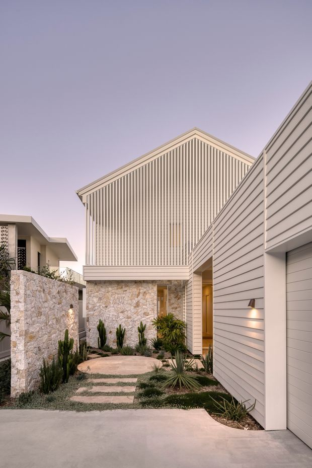A contemporary residential exterior viewed from the driveway, featuring a gabled upper volume with strong vertical screening, pale horizontal weatherboard-style cladding, natural irregular stone walls, and a landscaped entry garden with drought-tolerant planting and stepping stones. A garage occupies the right foreground, while neighbouring or adjoining low-profile structures are visible to the left.