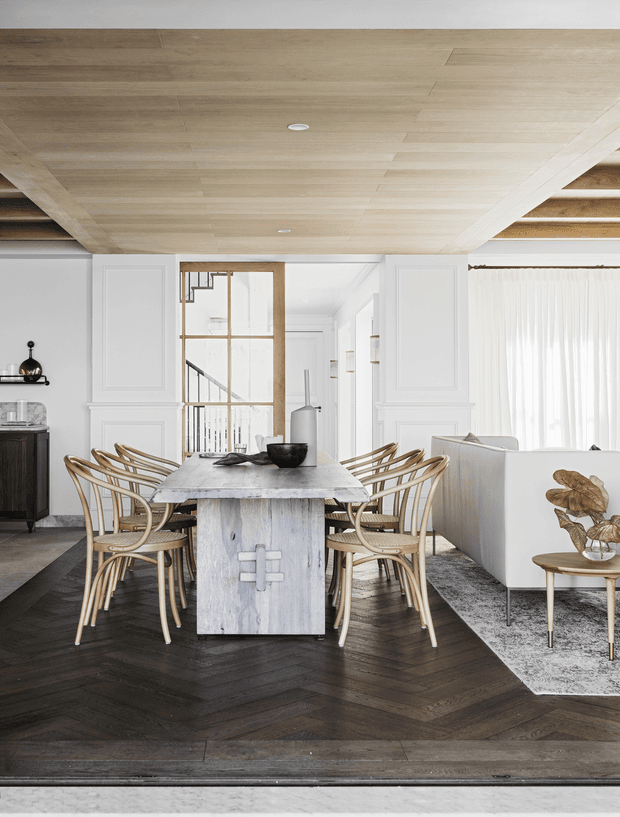 A refined open-plan dining area viewed from the living space, featuring a large pale stone dining table with sculptural pedestal base, curved woven timber dining chairs, dark herringbone flooring, white decorative wall panelling, and a light timber-lined ceiling. A glazed internal partition reveals a stairway and hallway beyond, while a white sofa and sheer curtains occupy the adjacent living area.