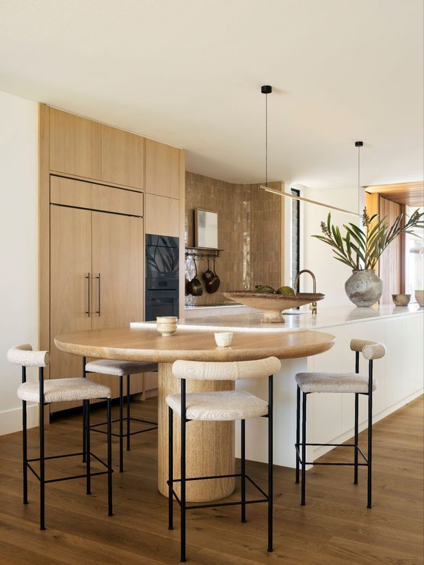 A warm, contemporary kitchen and dining area with a large central island, integrated light timber cabinetry and appliances, a rounded timber dining table, and upholstered black-framed bar stools. The island has a pale stone benchtop with a raised serving ledge, integrated sink and curved brass-toned tap. A textured brown tile feature wall, pendant lighting, decorative vessels, and timber flooring add warmth and texture.