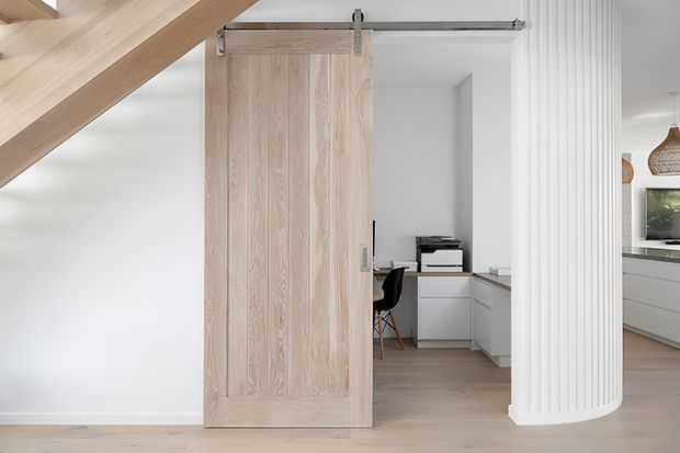 Bright contemporary interior beneath a staircase, featuring a large pale timber sliding barn door opening to a compact home office with a desk, printer and chair. A curved fluted white column separates the office from an adjacent kitchen or living area, with light timber flooring and white cabinetry throughout.