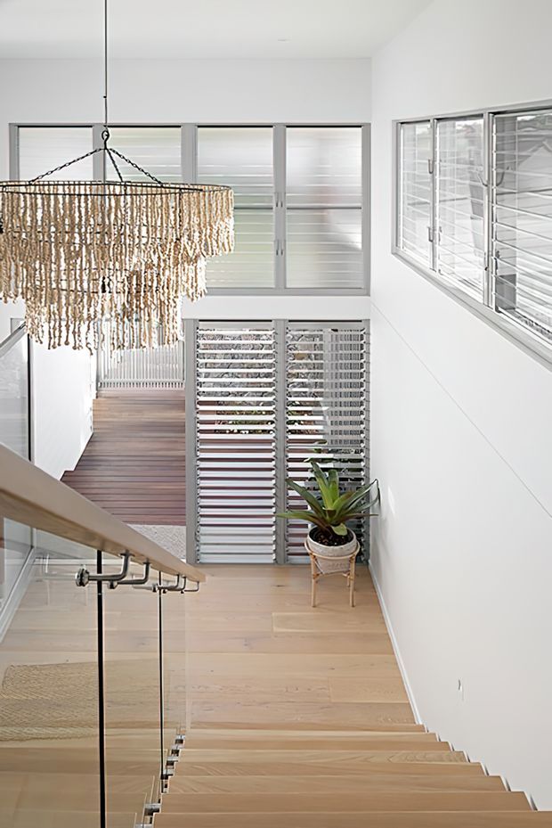Bright contemporary stair landing with pale timber stairs and flooring, white walls, frameless glass balustrades, and slim metal handrails. A large cascading beaded pendant hangs over the stairwell, while louvred screens and frosted glazing provide privacy and filtered light. A potted plant adds a natural accent.