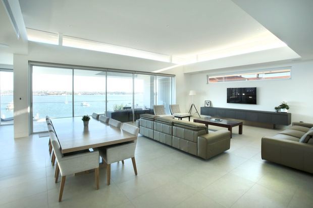 Bright, spacious contemporary open-plan living and dining area with floor-to-ceiling glazing overlooking a body of water. The room features pale tiled flooring, white walls and ceiling, recessed cove lighting, a large neutral sectional sofa, dining table with upholstered chairs, low timber media console, and minimalist furnishings.