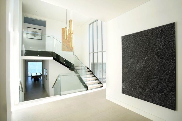 Bright contemporary multi-level entry and stair hall with a floating staircase, clear glass balustrades, dark contrasting stair treads and landing, tall vertical glazing, and sculptural gold pendant lights. A large dark textured wall artwork forms a feature surface on the right, while an open dining area is visible beyond the stair hall.