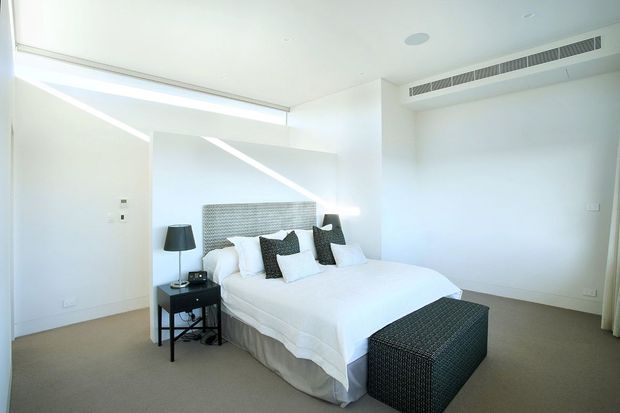 Bright, minimalist bedroom with a white upholstered bed, textured grey headboard, patterned dark cushions and matching upholstered bench. A black bedside table and lamp sit beside the bed, while concealed ceiling air-conditioning and broad angular window openings contribute to the clean contemporary design.