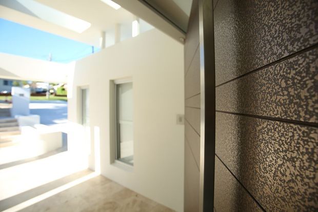 Close-up view of a contemporary residential entryway with a dark textured feature wall or cladding panel beside a recessed front door. The bright white facade, narrow vertical windows, covered porch, and clean geometric lines create a minimalist modern exterior.