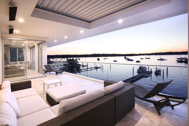 A luxurious covered outdoor terrace overlooking a calm waterfront marina, furnished with white upholstered sofas, dark woven lounge seating, and a low coffee table. The terrace features pale stone paving, recessed lighting, and a large louvred roof, with glazed doors opening into the interior.