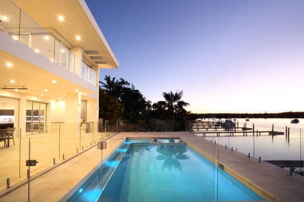 A modern waterfront residence at dusk, viewed from an elevated outdoor terrace. The image features a rectangular illuminated swimming pool enclosed by clear glass balustrades, a pale paved deck, expansive glazed living areas, a cantilevered upper level, and a calm marina or riverfront setting with private docks and palm trees.