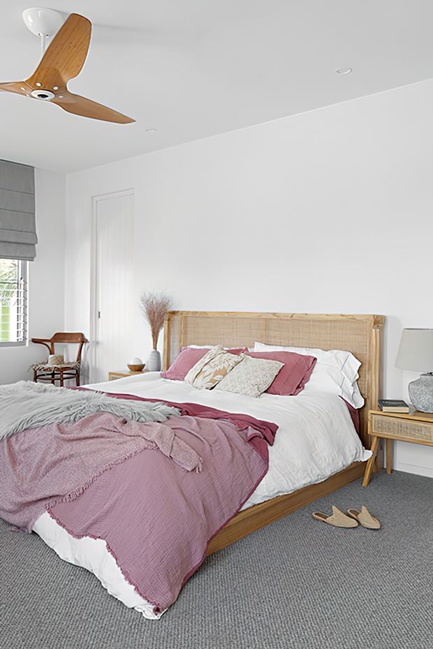 Bright, calm bedroom with a white-painted interior, grey carpet, and a timber bed featuring a woven cane/rattan headboard. The bed is dressed in white linens with dusty-rose and mauve throws and cushions. A timber ceiling fan, bedside table, window with Roman blind, armchair, and neutral accessories complete the relaxed contemporary coastal-inspired setting.