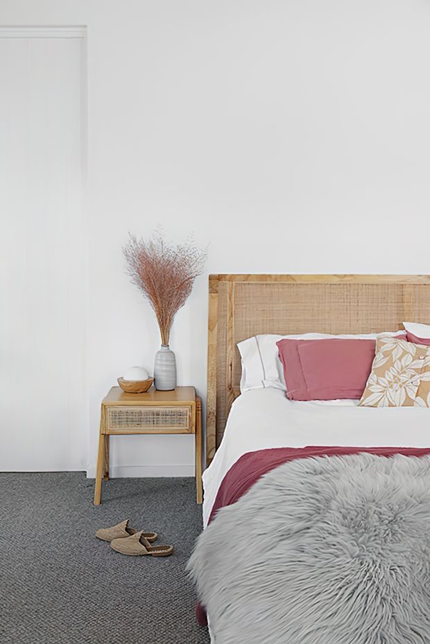 A bright, softly styled bedroom featuring a natural woven rattan headboard, white bedding, muted rose and floral accent pillows, and a large fluffy grey throw. A small timber bedside table with woven drawer fronts holds a vase of dried branches and a bowl. The room has white walls, grey carpet, and a calm, natural coastal-inspired atmosphere.