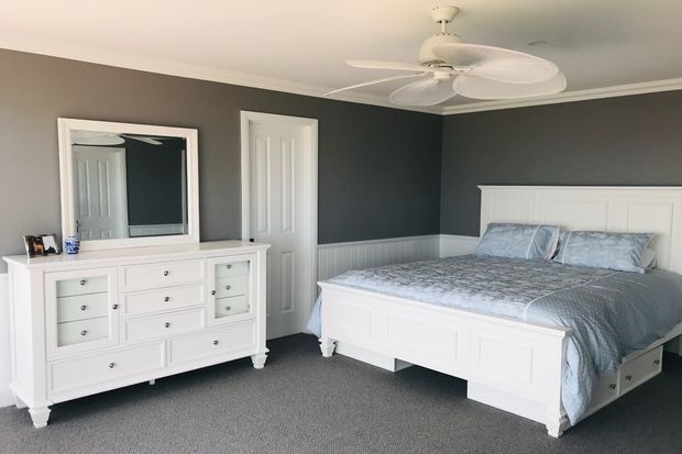 A bright, orderly bedroom with charcoal-grey walls, white wainscoting and trim, white panelled built-in-style furniture, and a large white panelled bed. A grey patterned duvet and pillows coordinate with the carpet, while a ceiling fan, dresser with mirror, and closed interior door complete the room.