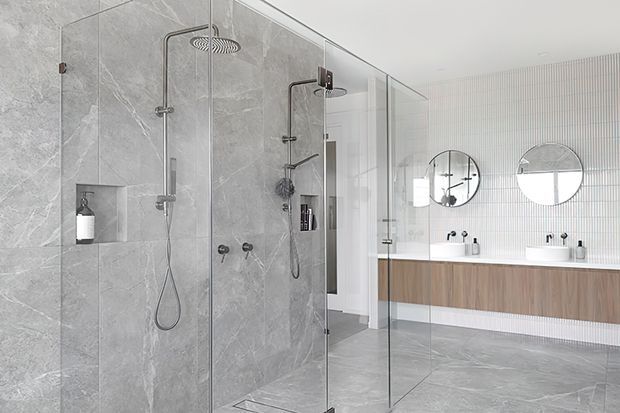 Contemporary bathroom featuring a large walk-in glass shower enclosure with dual rainfall showerheads, grey marble-look wall and floor tiles, and a floating timber vanity with twin white basins. A vertically ribbed light wall finish and round mirrors add texture and symmetry.