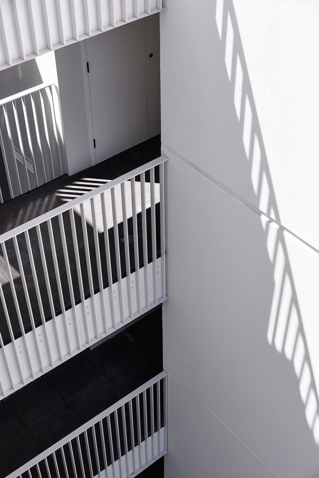 A stark, minimalist interior view looking down through a multi-level stairwell. White vertical balustrades and wall surfaces frame dark charcoal stair treads and landings, with a simple flush grey door at the upper landing. Strong sunlight casts repeating geometric railing shadows across the walls.