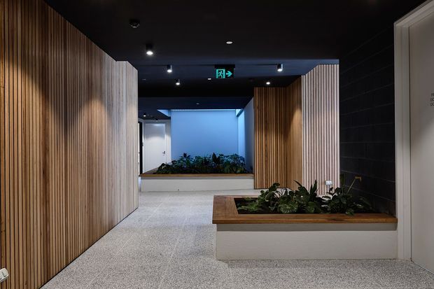 A contemporary commercial interior hallway or entry circulation space with dark ceilings, recessed spotlights, pale terrazzo-like flooring, and tall vertical timber slat wall panels. Built-in white planter boxes with integrated greenery define the corridor, while a blue-lit opening and illuminated exit sign are visible in the background.