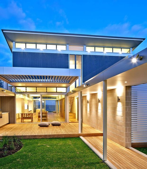A contemporary multi-level home arranged around a small landscaped courtyard. The exterior combines dark vertical cladding, pale rendered surfaces, extensive glazing, timber decking and a broad low-pitched roof with deep overhangs. A covered outdoor passage and carport-like structure frame the right side, while a pergola shades the glazed living and dining area. Warm evening lighting illuminates the timber deck, exposed masonry wall, stairs and courtyard.