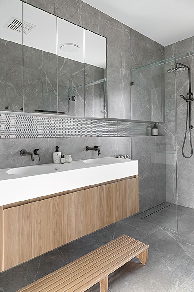 Contemporary bathroom featuring a floating double vanity with a white integrated benchtop and light timber cabinetry, large grey stone-look wall and floor tiles, a narrow hexagonal mosaic tile feature strip, frameless glass shower enclosure, black tapware and shower fittings, and a full-width mirrored wall cabinet. A timber slatted bench sits in the foreground.