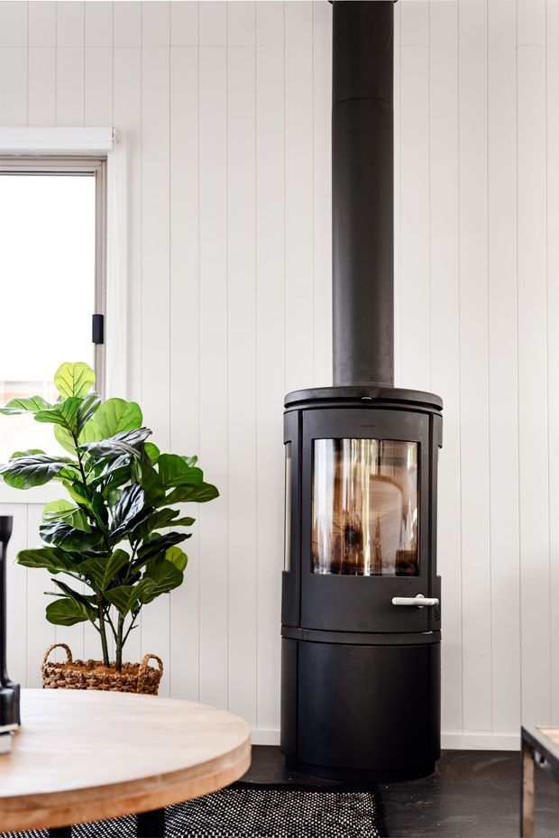 Bright interior corner featuring a freestanding black wood-burning stove with a tall cylindrical flue, set against white vertical timber wall panelling. A large leafy indoor plant sits beside a window, with a round timber table in the foreground and dark flooring below.