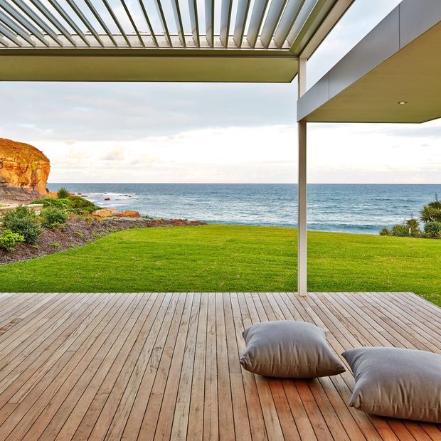 A modern open-air terrace overlooking the ocean, with a broad timber deck, green lawn, landscaped coastal garden and rocky shoreline. Slim structural posts support a large pergola with adjustable-looking metal louvres and a covered roof section, creating a sheltered outdoor lounge area. Two loose floor cushions sit on the deck. The scene is bright and relaxed, with strong coastal views and clean contemporary lines.