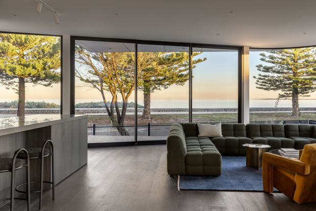 A contemporary open-plan living room with a large sectional sofa and armchair arranged around a low coffee table on a rug. Full-height glazed sliding doors and windows provide expansive views toward a waterfront or coastal landscape, while a minimalist kitchen island with bar stools occupies the left side. The space has dark-framed glazing, a smooth light ceiling with track lighting, and timber-look flooring.