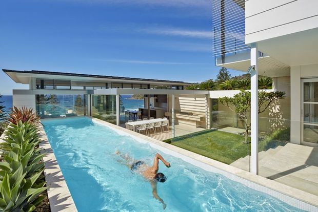 A bright contemporary coastal residence viewed from an elevated terrace, featuring a long narrow swimming pool in the foreground, landscaped planting, a covered outdoor dining area, expansive glass walls, and clean white architectural volumes. The ocean is visible beyond the property, with strong indoor-outdoor connections and abundant sunlight.