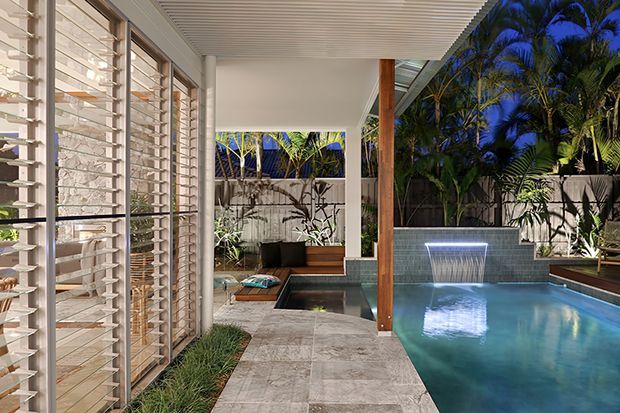 A covered outdoor pool courtyard beside a modern home, featuring a long tiled walkway, pale stone paving, timber posts, a slatted ceiling, louvred glazing, tropical planting, and a tiled pool with a small water feature. The space is illuminated at dusk with a calm, resort-like atmosphere.