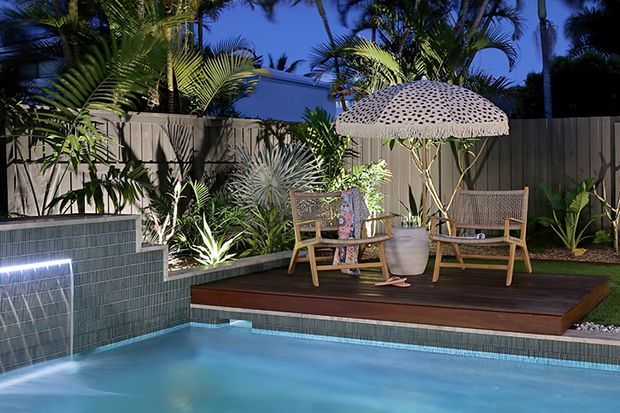 A tropical residential pool area at dusk, featuring a tiled swimming pool with illuminated water feature, a raised dark timber deck, woven outdoor lounge chairs, a decorative parasol, and dense palm and tropical planting enclosed by a timber fence.