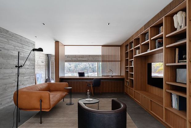 A refined home office and lounge with a built-in timber library wall, integrated desk, open shelving, and vertical timber screening around a large window. The room combines a warm caramel leather sofa and dark rounded lounge chair with a concrete-textured feature wall, dark flooring, and minimalist black lighting. A forested outlook is visible through the glazing.