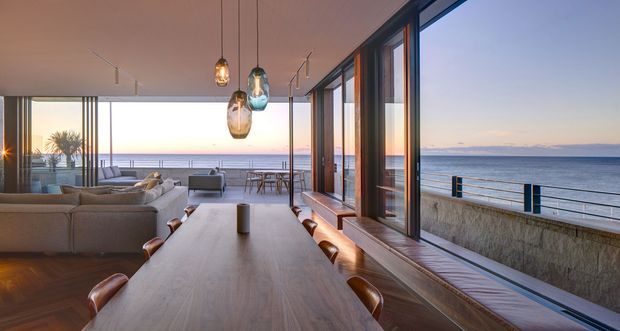 A refined open-plan coastal living and dining space with expansive floor-to-ceiling glazing overlooking the ocean. A long timber dining table with leather chairs occupies the foreground, while a large neutral sectional lounge sits beyond it. Warm pendant lights, timber flooring, dark-framed glazing, and built-in window seating create a calm, sunset-lit interior with a direct connection to the waterfront terrace.