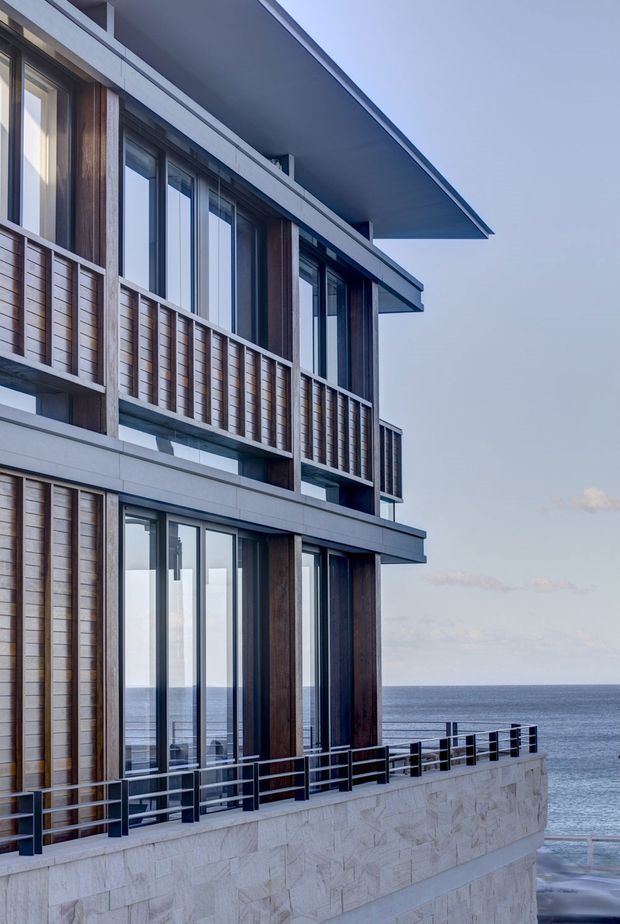 Exterior architectural view of a multi-level modern coastal residence overlooking the ocean. The facade combines pale stone or concrete-like wall bands, dark timber vertical cladding, extensive floor-to-ceiling glazing, projecting flat roof planes, and metal balcony railings. A recessed balcony and ocean-facing terraces are visible, with the sea forming the background.