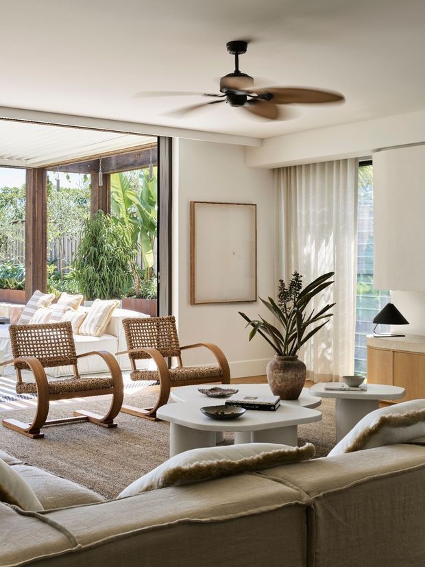 Bright, warm living room with a large cream sectional sofa in the foreground, two woven wood lounge chairs, rounded white coffee tables, a timber media console, a potted plant, and sheer curtains. The room opens to a leafy outdoor sitting area through extensive glazing, with a ceiling fan and soft neutral finishes creating an airy contemporary interior.