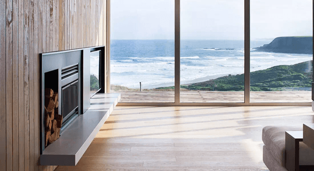 A minimalist coastal living room with a built-in linear fireplace set into vertical timber wall panelling. Full-height glazed doors or windows open to expansive ocean and coastal headland views, while warm timber flooring and restrained furnishings create a bright, serene interior.