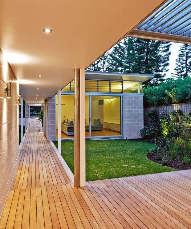 A contemporary residential courtyard viewed from a covered timber deck and walkway. The space features warm timber decking, a flat covered roof with recessed downlights, slender timber posts, extensive glazing, pale grey masonry walls, and a small lawn bordered by established planting. A compact living area is visible through the glass, while a glazed roof section brings additional daylight into the covered outdoor space.