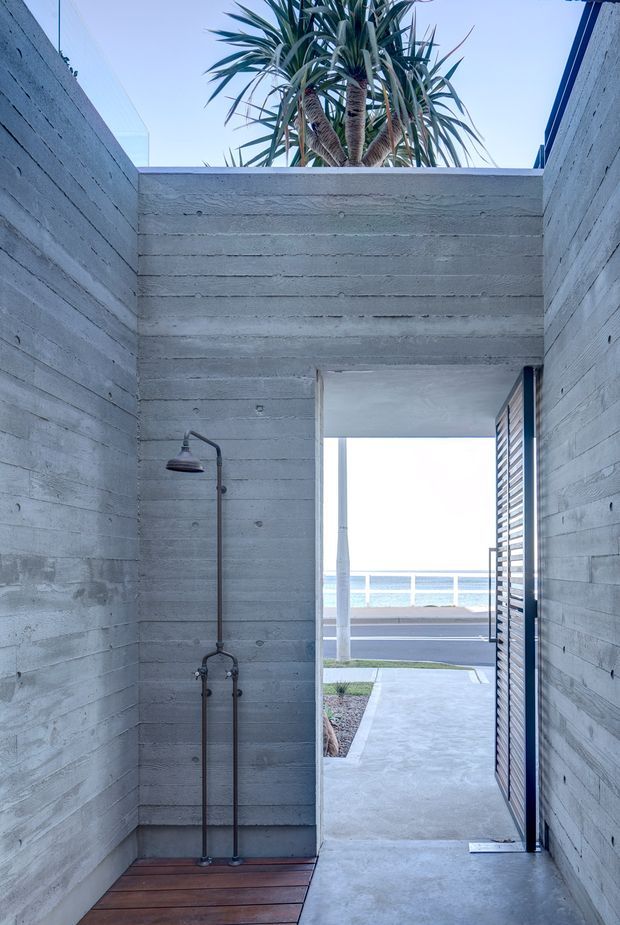 A narrow open-air architectural passage with an outdoor shower, enclosed by horizontally board-formed concrete walls and a concrete overhead beam. A small timber deck sits beneath the dark metal shower fixture, while a louvred door opens toward a paved exterior path, road, and waterfront railing. Tropical sculptural planting rises above the wall, creating a stark coastal modern composition.