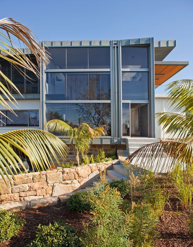 Exterior view of a contemporary multi-storey home with extensive floor-to-ceiling glazing, a strongly articulated grey metal-framed facade, projecting roof canopies, and a recessed entry. Tropical palms and native-style landscaping surround the building, with a low natural stone retaining wall and stepped path in the foreground.