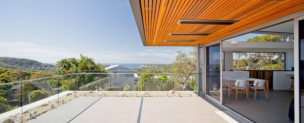 A bright contemporary residential terrace overlooking a leafy coastal or hilly landscape. The expansive outdoor area has pale concrete or stone paving, frameless glass balustrades, and a dramatic timber-lined projecting ceiling with slim recessed linear lights. Full-height glazed sliding doors open into a modern dining and kitchen area, with white cabinetry, a dark island or benchtop, and light timber dining chairs visible inside.