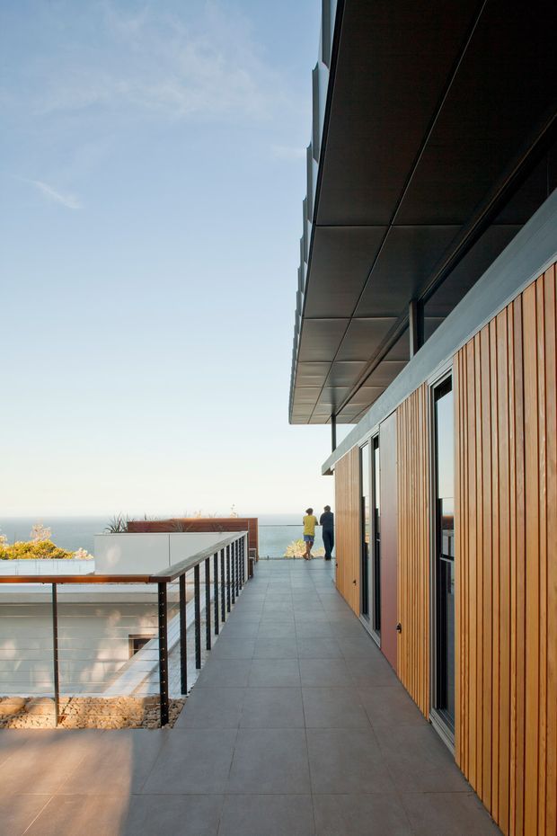 A modern coastal exterior walkway or terrace running alongside a building, with warm vertical timber cladding, dark metal-framed glazing, a broad projecting roof canopy, and large grey tiled paving. A glass-and-metal balustrade overlooks the ocean, with a distant horizon and landscaped planting visible beyond.