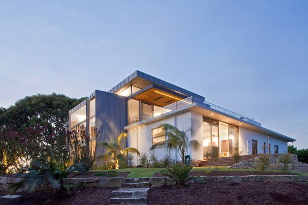 Twilight exterior view of a contemporary two-storey residence on a landscaped, slightly elevated site. The house features crisp white rendered walls, extensive floor-to-ceiling glazing, projecting flat roof planes, dark metal cladding, warm timber soffits, illuminated balconies, and broad outdoor steps. Tropical palms and low planting surround the modern facade.