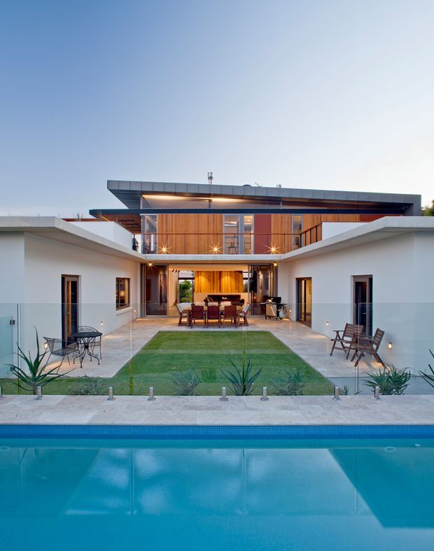 Symmetrical view of a contemporary two-storey courtyard house from across a swimming pool. White rendered wings frame a central outdoor dining and entertaining area, with warm timber wall cladding and extensive glazing on the upper level. The home has broad flat roof planes, a central lawn, minimalist landscaping, and a calm blue evening sky.