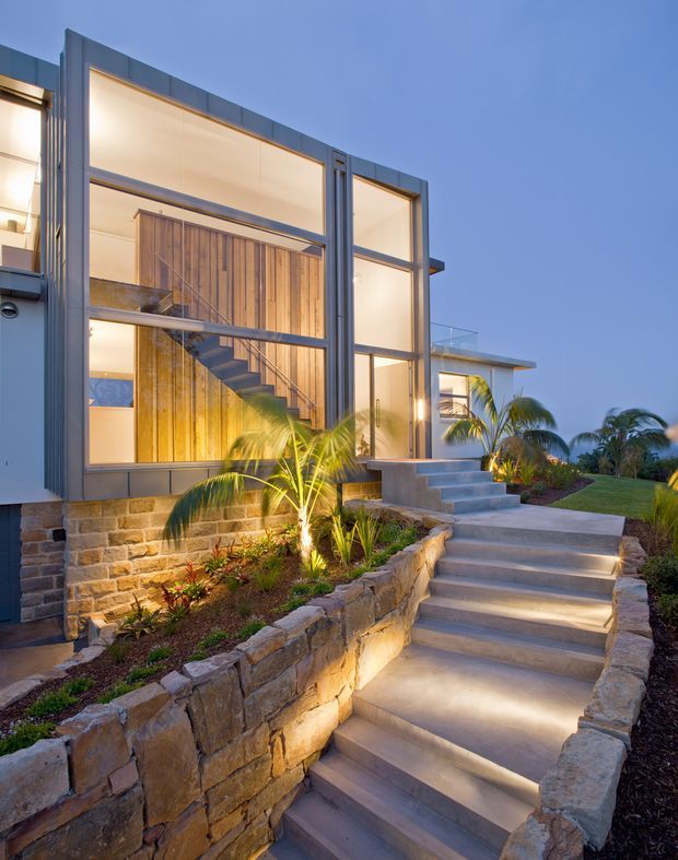 Twilight exterior view of a contemporary multi-level residence with extensive floor-to-ceiling glazing, dark metal framing, and a prominent internal timber staircase visible through the windows. Broad illuminated concrete steps lead through landscaped planting and natural stone retaining walls toward the entrance. Palm trees and warm architectural lighting create a refined resort-like atmosphere.