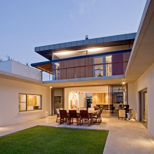 Twilight view of a contemporary multi-storey courtyard residence with a flat roof, projecting upper-level volumes, extensive glazing, timber screening, and an open-plan ground-floor entertaining area. A dining table and outdoor kitchen or barbecue occupy the paved courtyard beside a small lawn, while warm interior lighting creates an inviting indoor-outdoor connection.