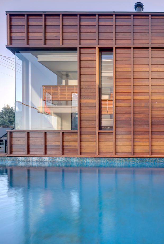 Close exterior view of a contemporary multi-storey home beside a swimming pool. The facade is clad in warm horizontal timber boards with regularly spaced vertical timber battens, large floor-to-ceiling glazing, and a projecting upper level. Interior spaces and a timber balcony are visible through the glass, while the pool occupies the foreground.