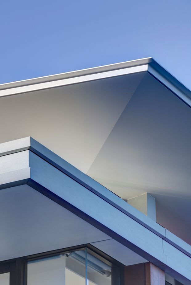 Close-up architectural photograph of a contemporary building corner and roof/eave junction, showing crisp angular planes, projecting horizontal fascia elements, smooth pale wall surfaces, dark metal detailing, and a glazed corner below. The composition is minimal and geometric against a clear blue sky.