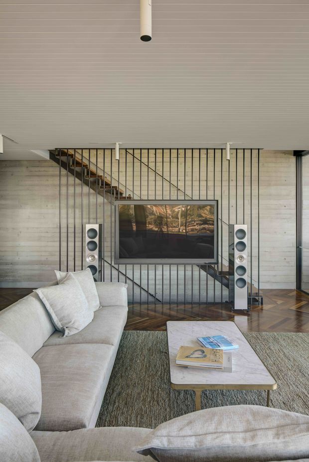 A contemporary living room with a large pale upholstered sectional sofa, low rectangular coffee table, and floor-standing speakers flanking a wall-mounted television. The room features a striking timber parquet floor, light horizontal timber wall and ceiling lining, and a black metal stair balustrade behind the media area, creating a warm modern aesthetic.