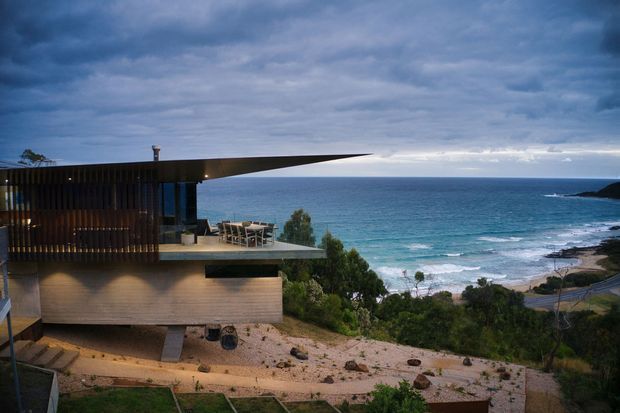 A modern coastal house perched on a sloping site above the ocean. The building has a dramatic low, cantilevered roof projecting toward the water, dark vertical timber screening, expansive glazing, and a projecting outdoor terrace with dining furniture. The surrounding landscape is rugged and sparsely planted, with a beach and rocky shoreline visible beyond. The image has a moody, dramatic atmosphere under an overcast sky.