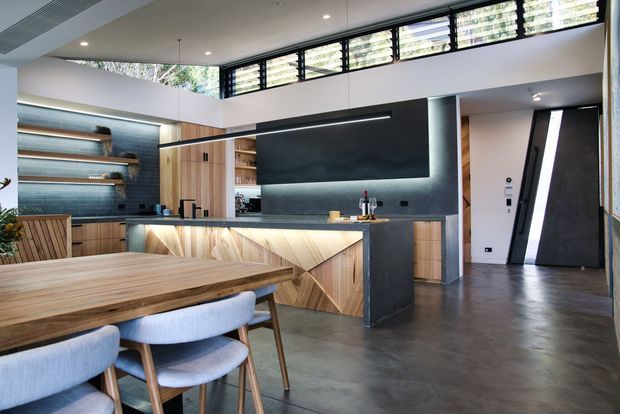 Open-plan contemporary kitchen and dining area with a large timber dining table in the foreground, pale upholstered dining chairs, timber kitchen cabinetry, a sculptural illuminated island, dark textured splashback and feature wall, and high-level louvred windows admitting natural light. The room has polished concrete flooring, a high ceiling, and a partly visible entry area.