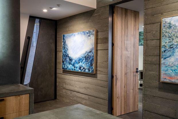 A moody contemporary interior entry or hallway with dark horizontally textured wall panels, a tall narrow glazed door or window, a timber pivot-style door, and a dark stone or concrete-like console. Blue-toned artwork adds contrast against the restrained grey-brown palette.